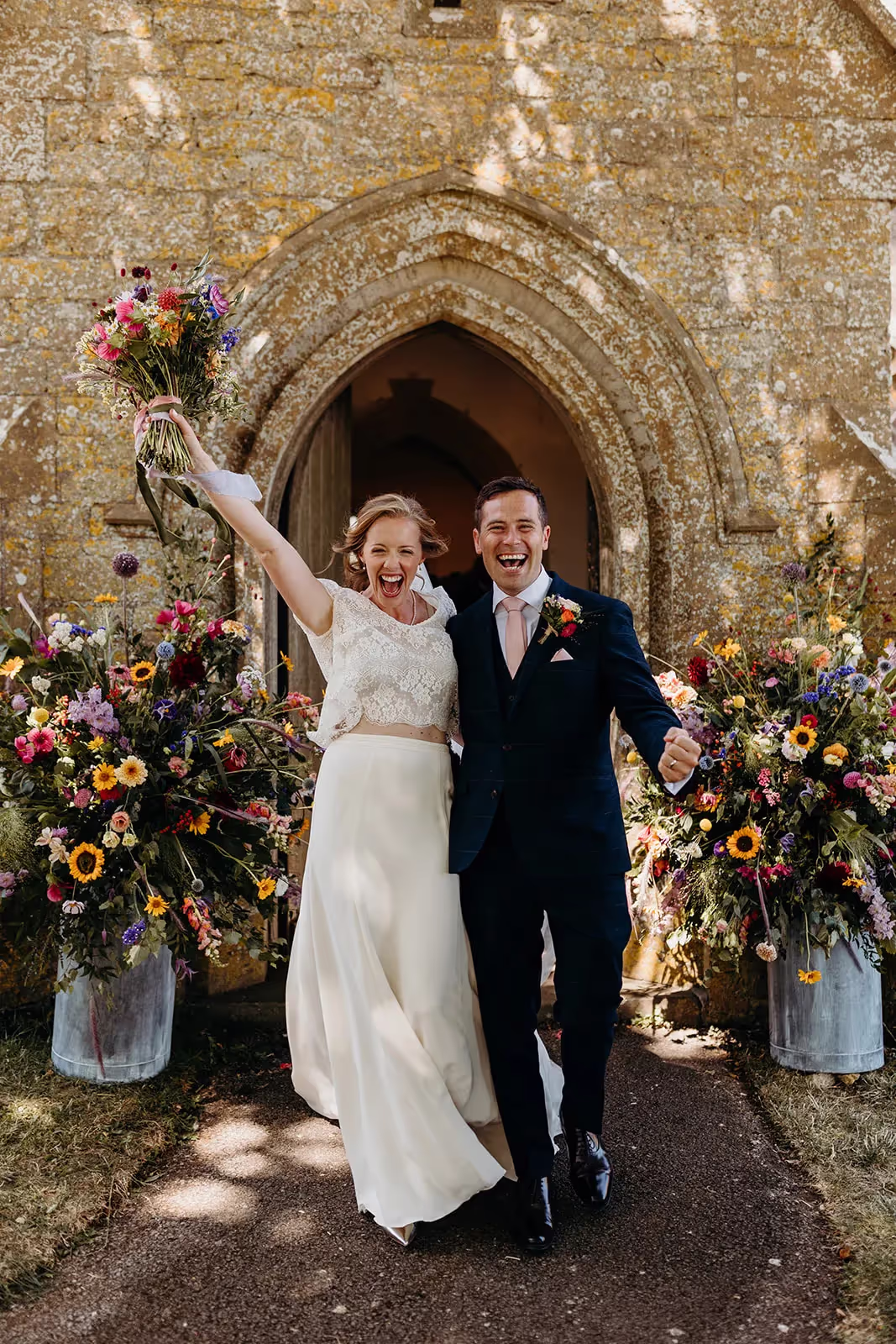Bride and groom leaving church with large flowering displays either side, bride holds bouquet in air with beaming smile