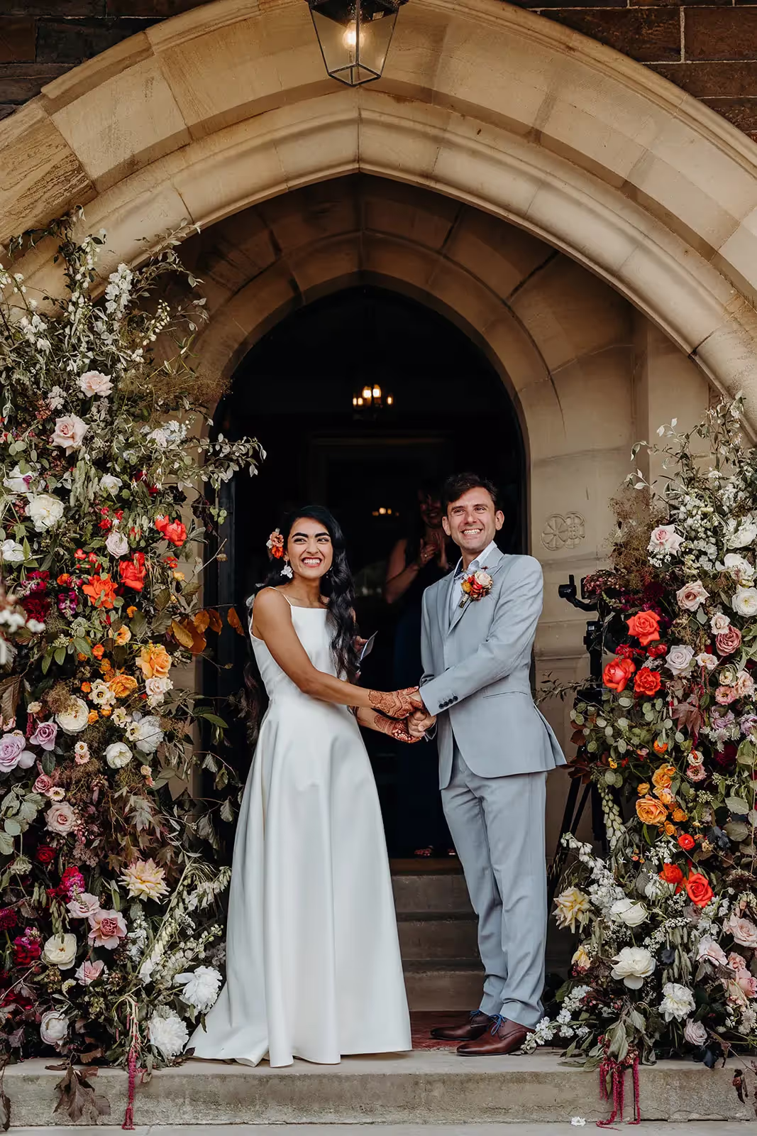 Outdoor ceremony at entrance of country house with bride and groom holding hands and floral displays either side